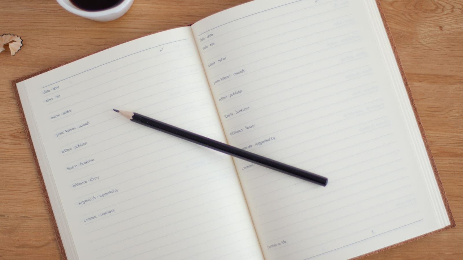 An open notebook with a pencil next to a cup of coffee on a wooden desk