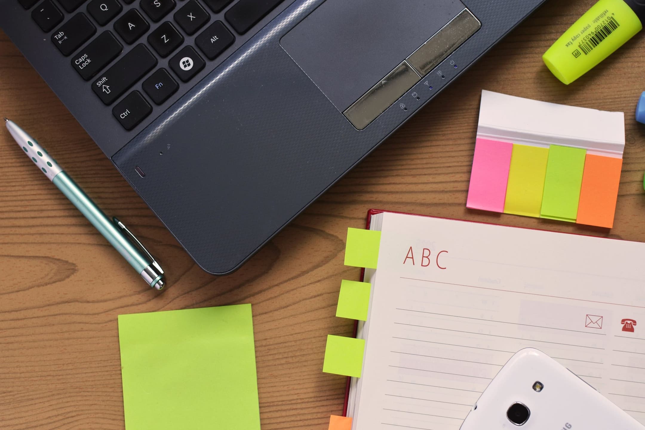 A laptop, a notebook, a pen and colourful sticky notes on a wooden desk